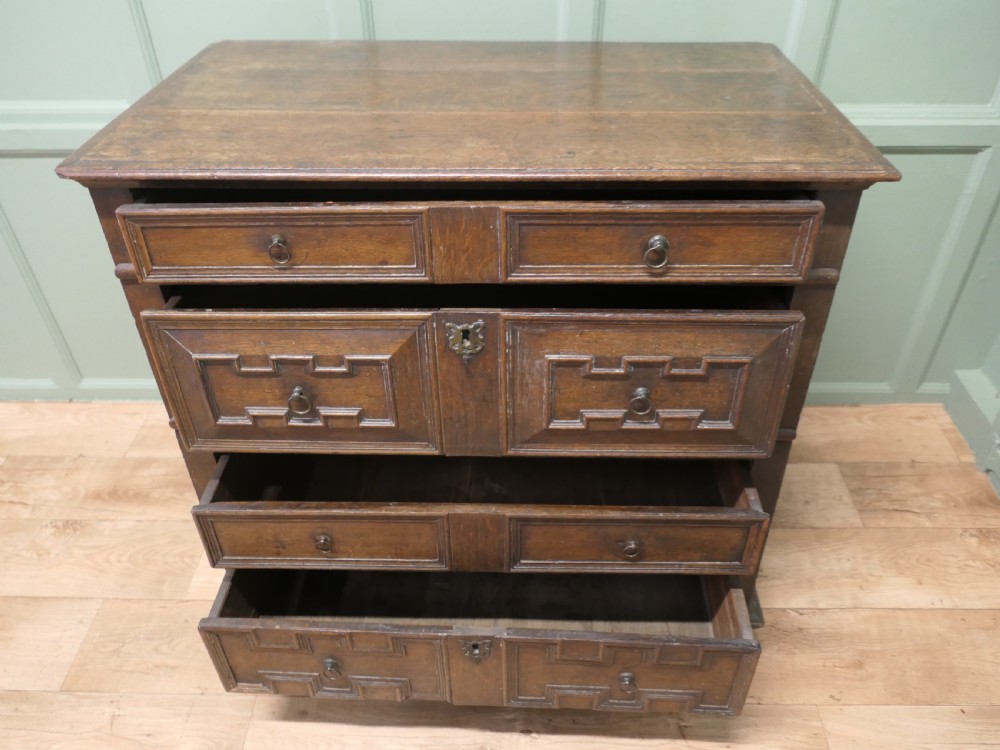 An 18th century dark oak chest of drawers featuring geometric panelled drawer fronts, brass drop handles, and panelled sides. View V