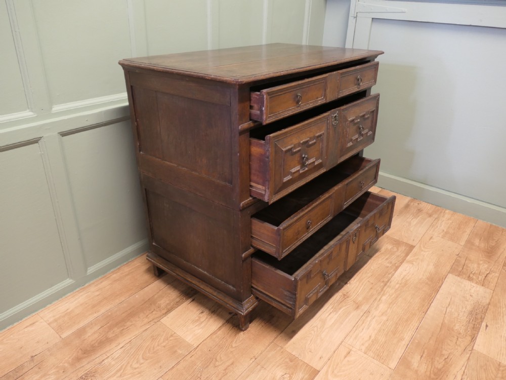 An 18th century dark oak chest of drawers featuring geometric panelled drawer fronts, brass drop handles, and panelled sides. View VI