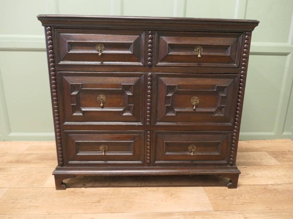 A dark oak chest of drawers featuring geometric moulded panels, brass teardrop handles, and vertical bobbin moulding.