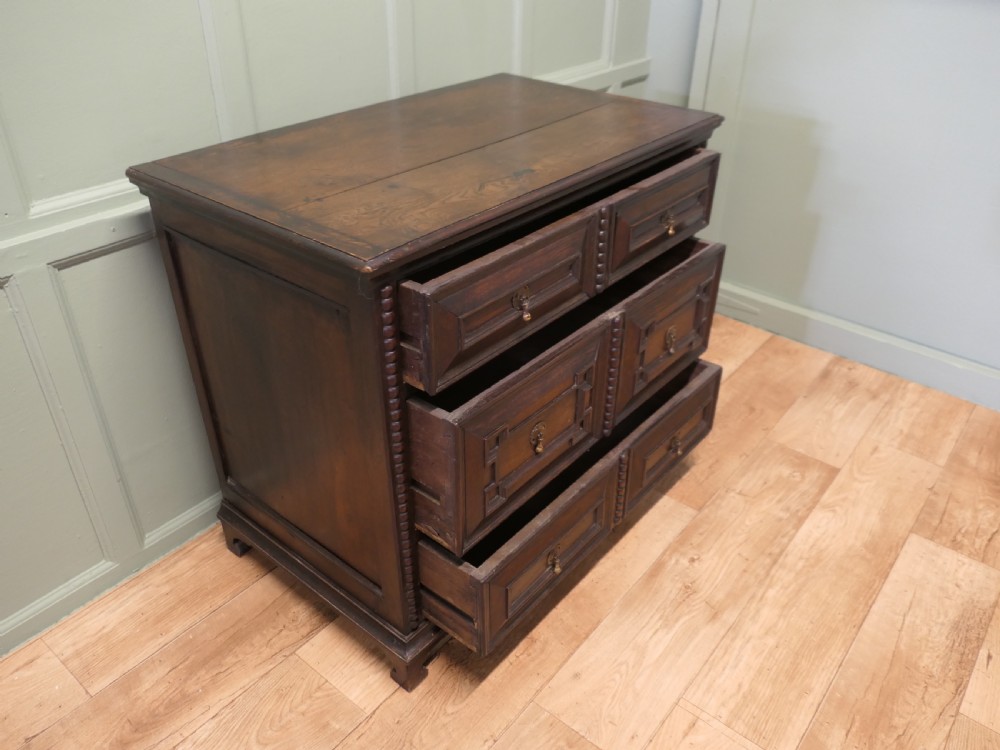 A dark oak chest of drawers featuring geometric moulded panels, brass teardrop handles, and vertical bobbin moulding. View II