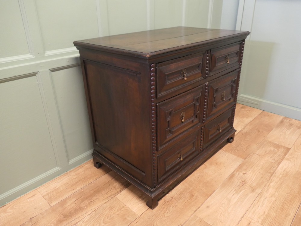 A dark oak chest of drawers featuring geometric moulded panels, brass teardrop handles, and vertical bobbin moulding. View III