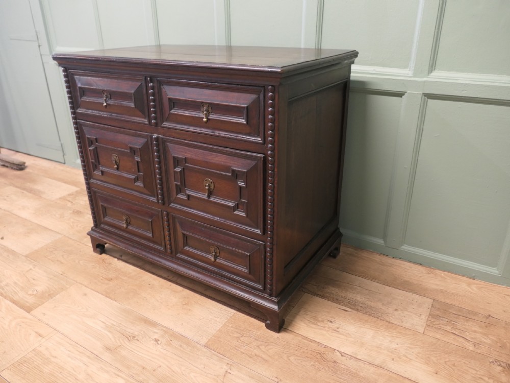 A dark oak chest of drawers featuring geometric moulded panels, brass teardrop handles, and vertical bobbin moulding. View V