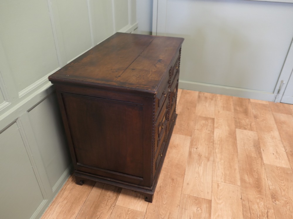 A dark oak chest of drawers featuring geometric moulded panels, brass teardrop handles, and vertical bobbin moulding. View VI