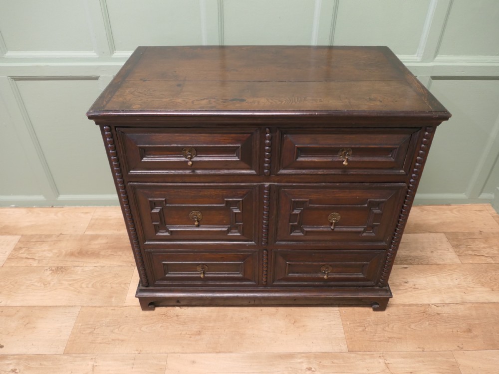 A dark oak chest of drawers featuring geometric moulded panels, brass teardrop handles, and vertical bobbin moulding. View VII