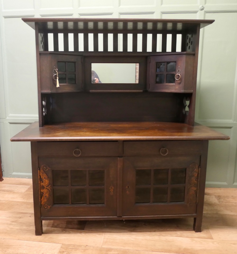 An Arts and Crafts dark oak sideboard featuring a slatted upper gallery, central mirror, two small upper cabinets, frieze drawers, and lower cupboards with strap hinges.