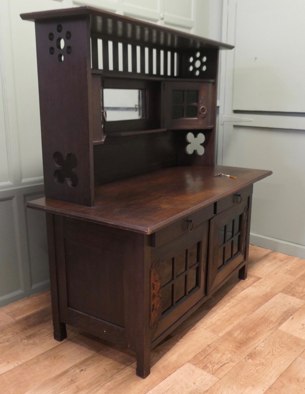 An Arts and Crafts dark oak sideboard featuring a slatted upper gallery, central mirror, two small upper cabinets, frieze drawers, and lower cupboards with strap hinges. View II