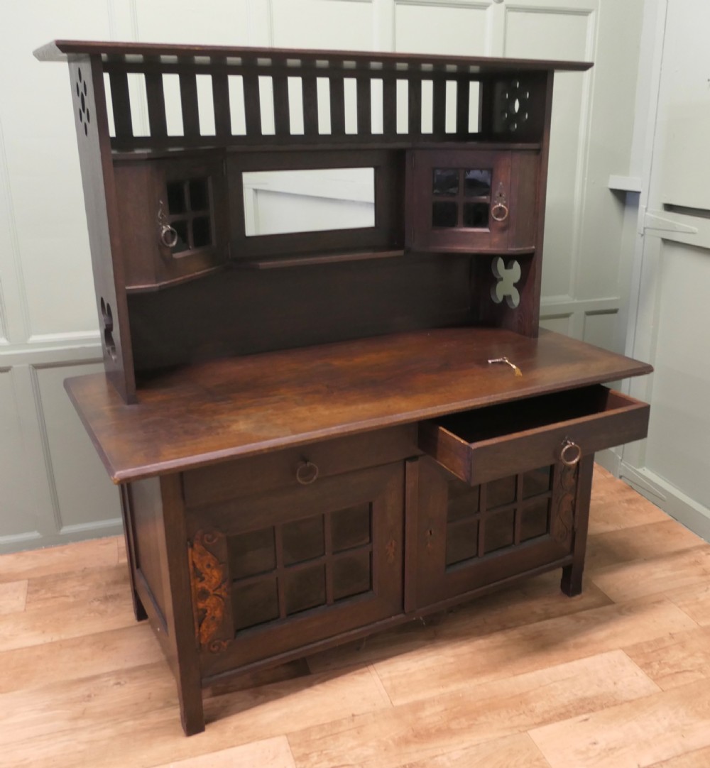 An Arts and Crafts dark oak sideboard featuring a slatted upper gallery, central mirror, two small upper cabinets, frieze drawers, and lower cupboards with strap hinges. View IV