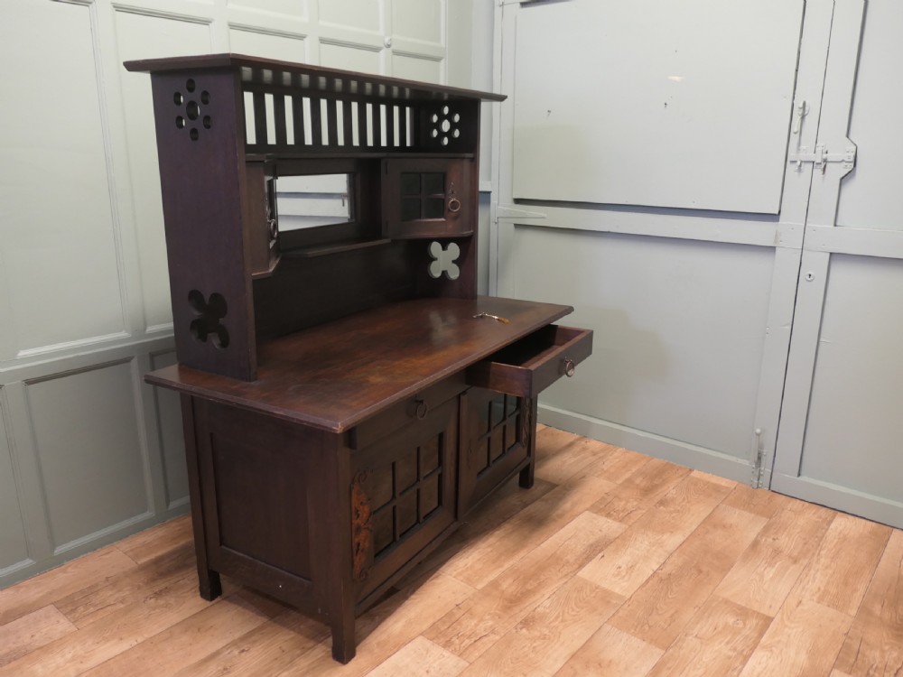 An Arts and Crafts dark oak sideboard featuring a slatted upper gallery, central mirror, two small upper cabinets, frieze drawers, and lower cupboards with strap hinges. View V