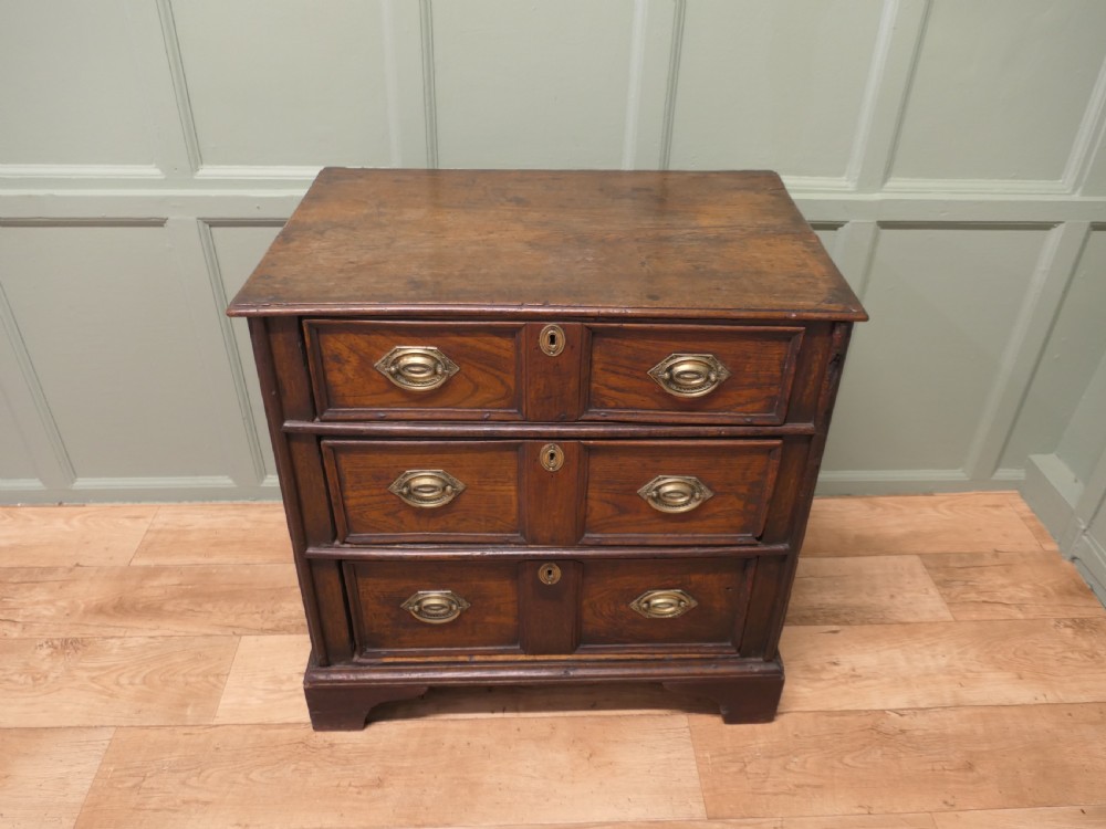 An 18th-century country elm chest of drawers with three moulded drawer fronts, brass drop handles, keyhole escutcheons, and bracket feet.