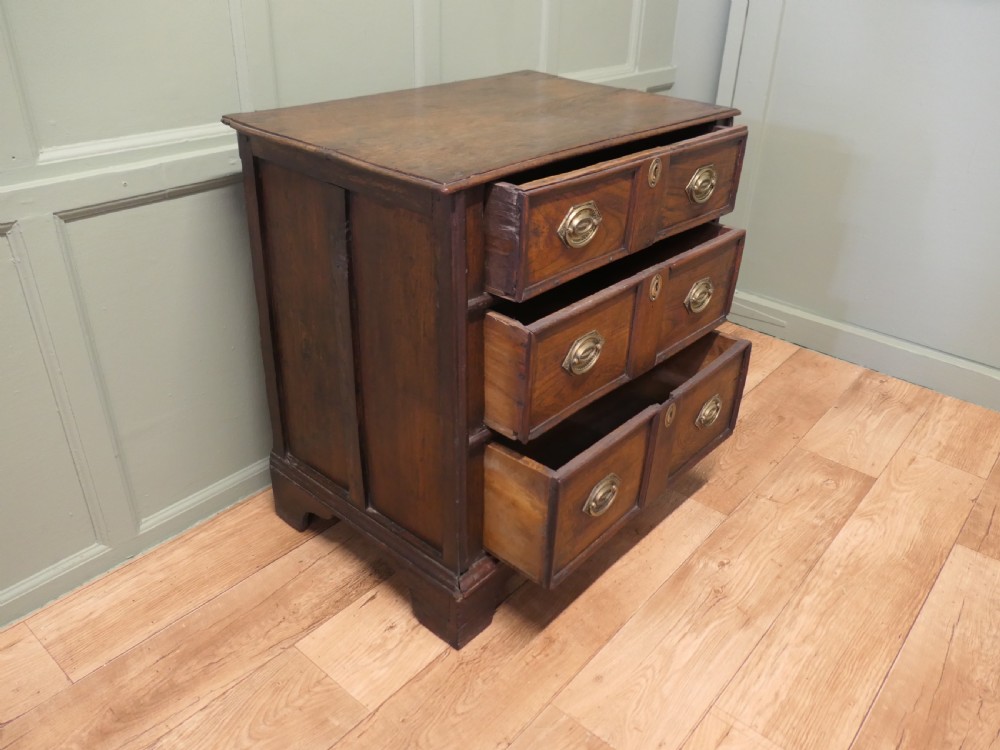 An 18th-century country elm chest of drawers with three moulded drawer fronts, brass drop handles, keyhole escutcheons, and bracket feet. View IV