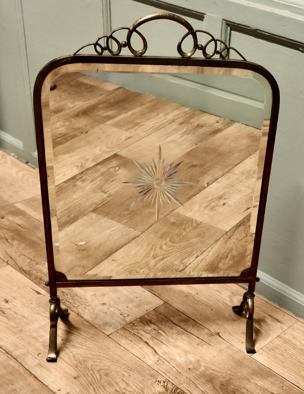 An Art Deco brass fire screen featuring a bevelled glass mirror panel with a cut sunburst motif set on cast metal feet.