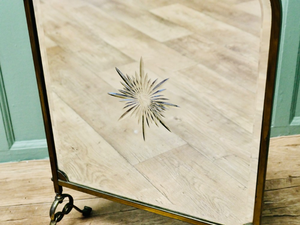 An Art Deco brass fire screen featuring a bevelled glass mirror panel with a cut sunburst motif set on cast metal feet. View V