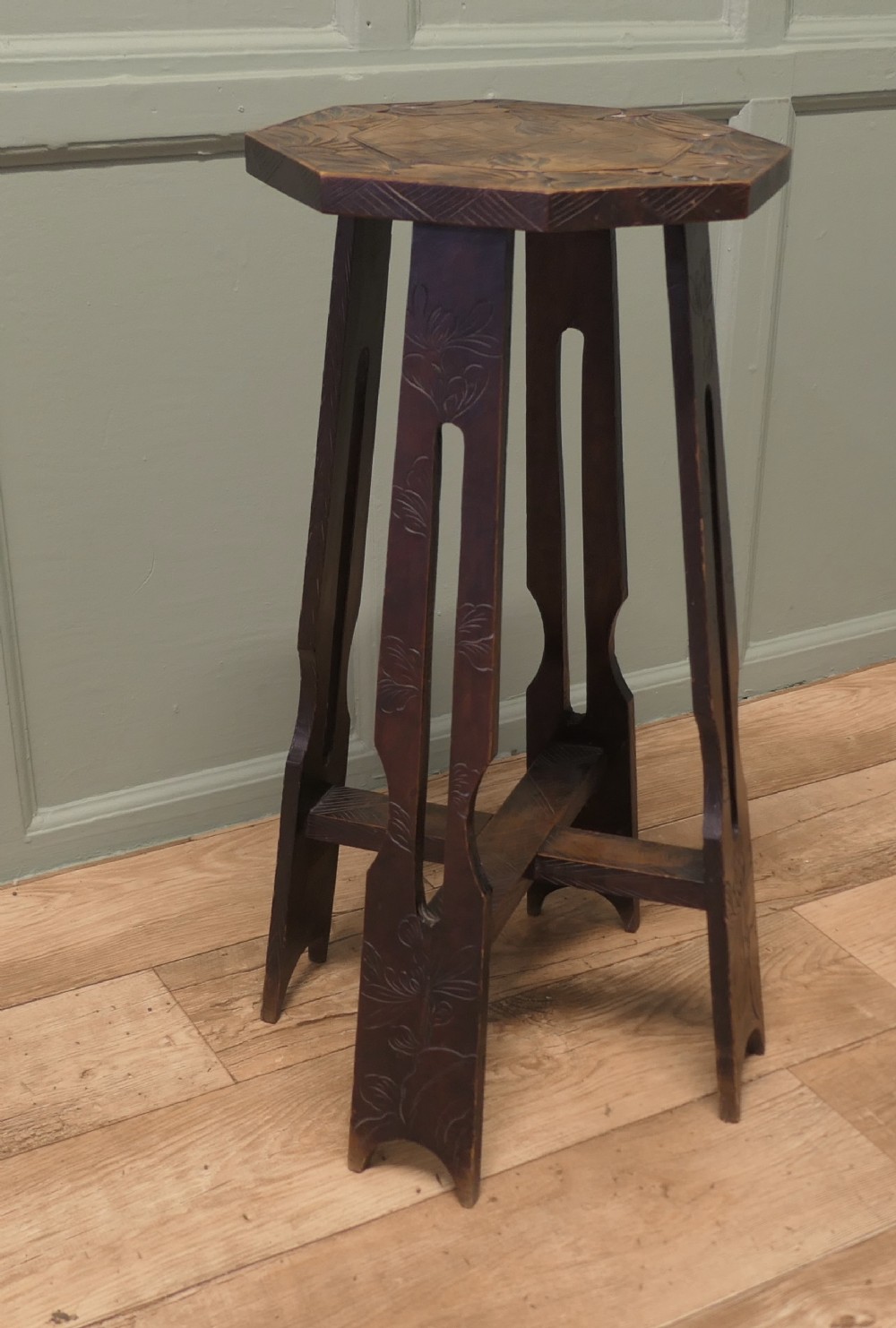 An Arts and Crafts dark elm plant stand featuring an octagonal carved top, stylised floral carving on four pierced legs, and an X-frame stretcher.