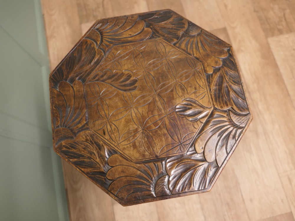 An Arts and Crafts dark elm plant stand featuring an octagonal carved top, stylised floral carving on four pierced legs, and an X-frame stretcher. View IV