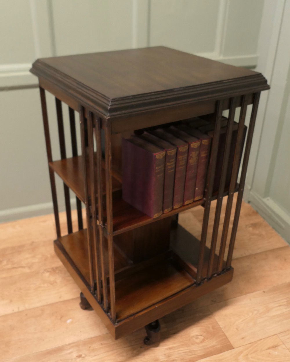 A square mahogany revolving library bookcase featuring vertical slatted sides, three tiered shelves, and castors on the base.
