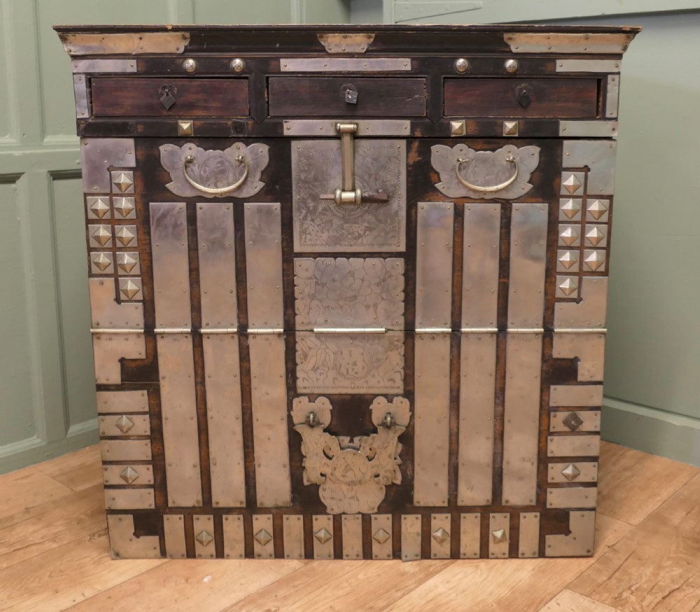 A dark brown wooden Korean Bandaji chest adorned with elaborate silver-coloured brass plating, geometric studs, etched metal panels, and three top drawers.