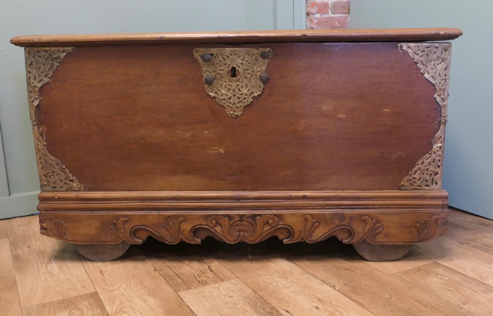 A rectangular carved elm wood storage coffer featuring decorative pierced brass corner mounts and a central lockplate above an ornate carved apron.