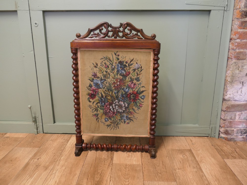 A Victorian mahogany fire screen featuring spiral barley twist supports, an openwork carved scroll cresting, and a central needlework panel depicting a colourful floral bouquet.