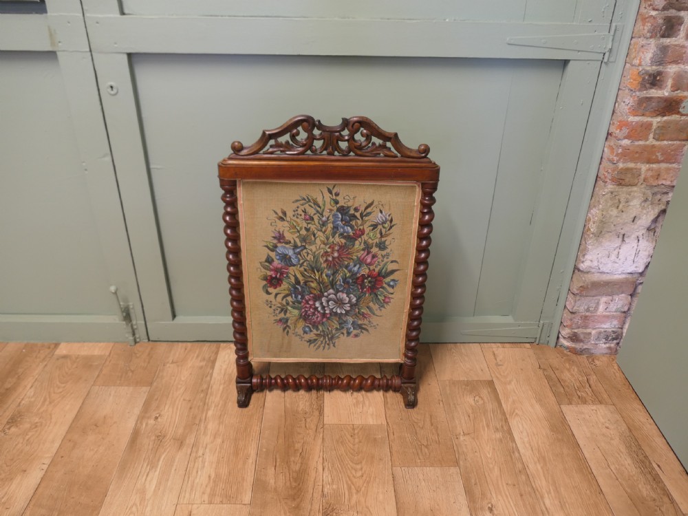 A Victorian mahogany fire screen featuring spiral barley twist supports, an openwork carved scroll cresting, and a central needlework panel depicting a colourful floral bouquet. View II