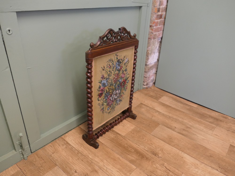 A Victorian mahogany fire screen featuring spiral barley twist supports, an openwork carved scroll cresting, and a central needlework panel depicting a colourful floral bouquet. View III