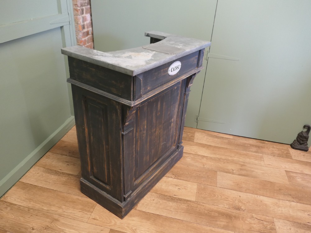 A distressed dark charcoal painted timber greeting desk with a zinc surface and an enamel plaque inscribed with the word Caisse.