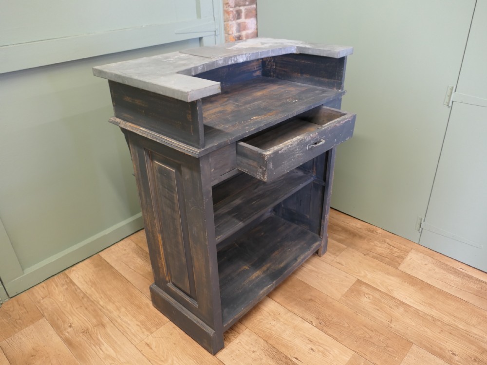 A distressed dark charcoal painted timber greeting desk with a zinc surface and an enamel plaque inscribed with the word Caisse. View IV