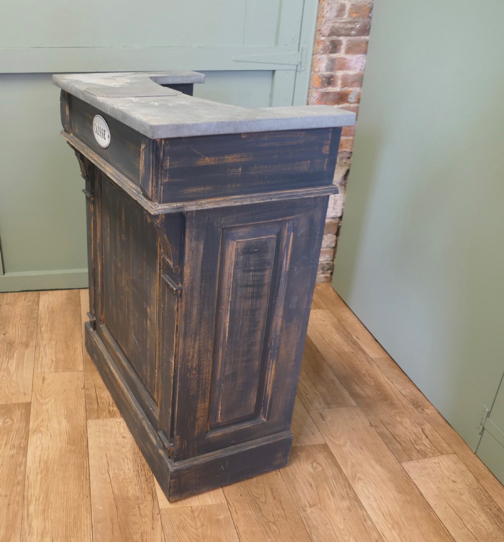 A distressed dark charcoal painted timber greeting desk with a zinc surface and an enamel plaque inscribed with the word Caisse. View V