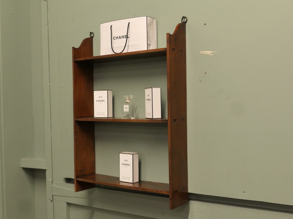 A three-tier open wooden wall shelf unit with a warm brown finish and metal hanging rings at the top. View II