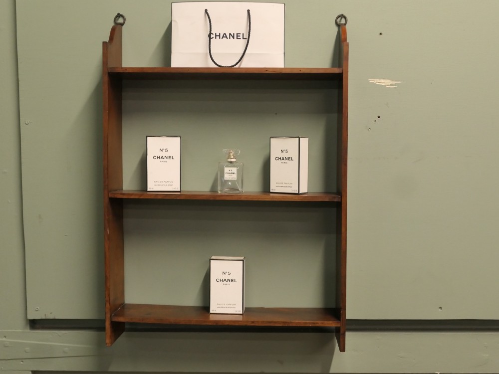 A three-tier open wooden wall shelf unit with a warm brown finish and metal hanging rings at the top. View III