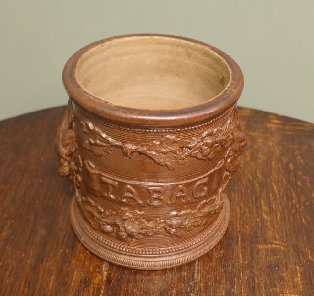 A cylindrical brown glazed stoneware tobacco pot featuring raised lettering that reads TABAC surrounded by oak leaf and acorn garlands.