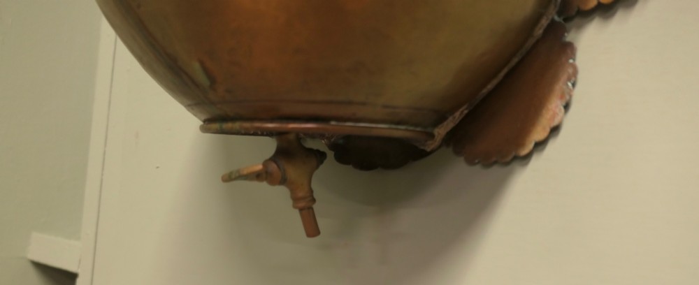 A hand-crafted copper and brass wall-mounted water reservoir featuring a scalloped backplate, rope-twist waist band, and lower tap. View V