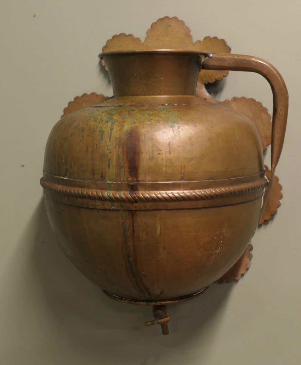 A hand-crafted copper and brass wall-mounted water reservoir featuring a scalloped backplate, rope-twist waist band, and lower tap. View VI