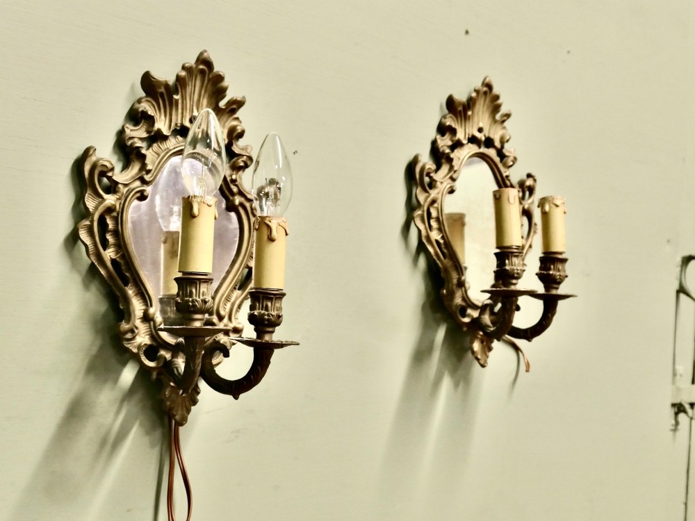 A pair of ornate gilt brass Rococo wall sconces featuring oval central mirrors and twin candle-style light fittings.