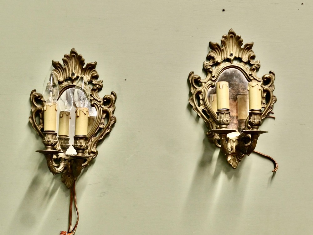 A pair of ornate gilt brass Rococo wall sconces featuring oval central mirrors and twin candle-style light fittings. View IV