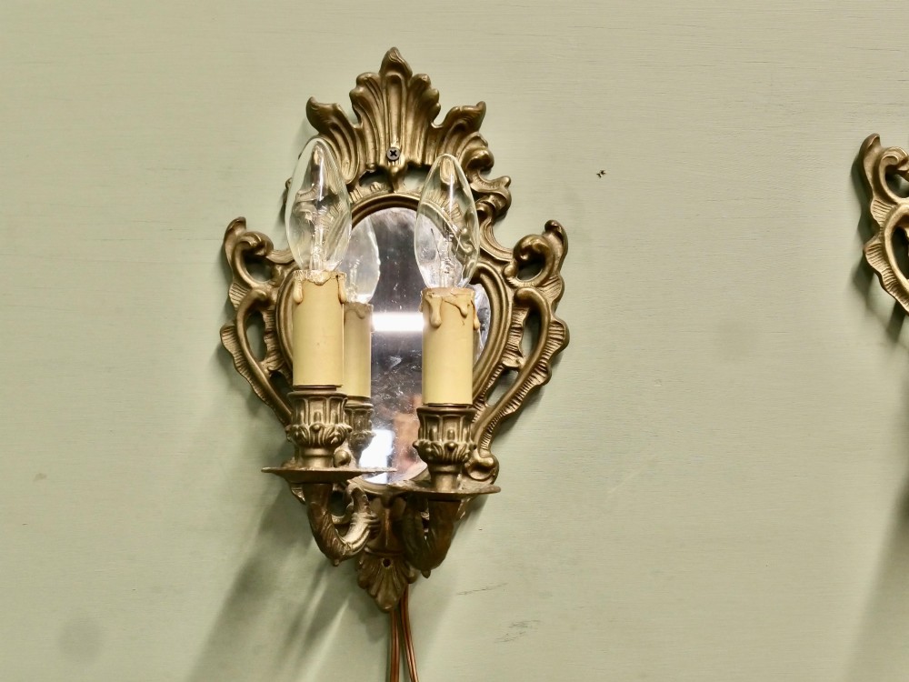 A pair of ornate gilt brass Rococo wall sconces featuring oval central mirrors and twin candle-style light fittings. View V