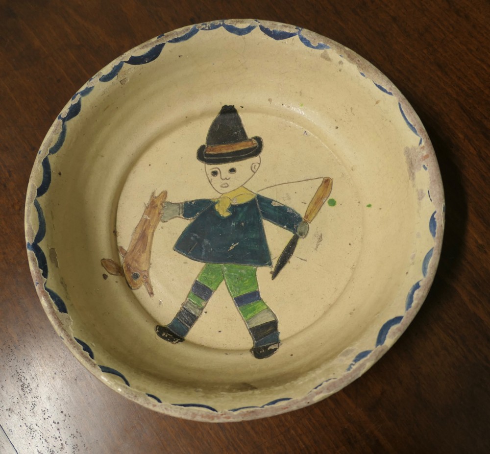 A large round cream-coloured French slipware terracotta bowl decorated with a blue scalloped rim and a naive central illustration of a hunter carrying a bow and hare.