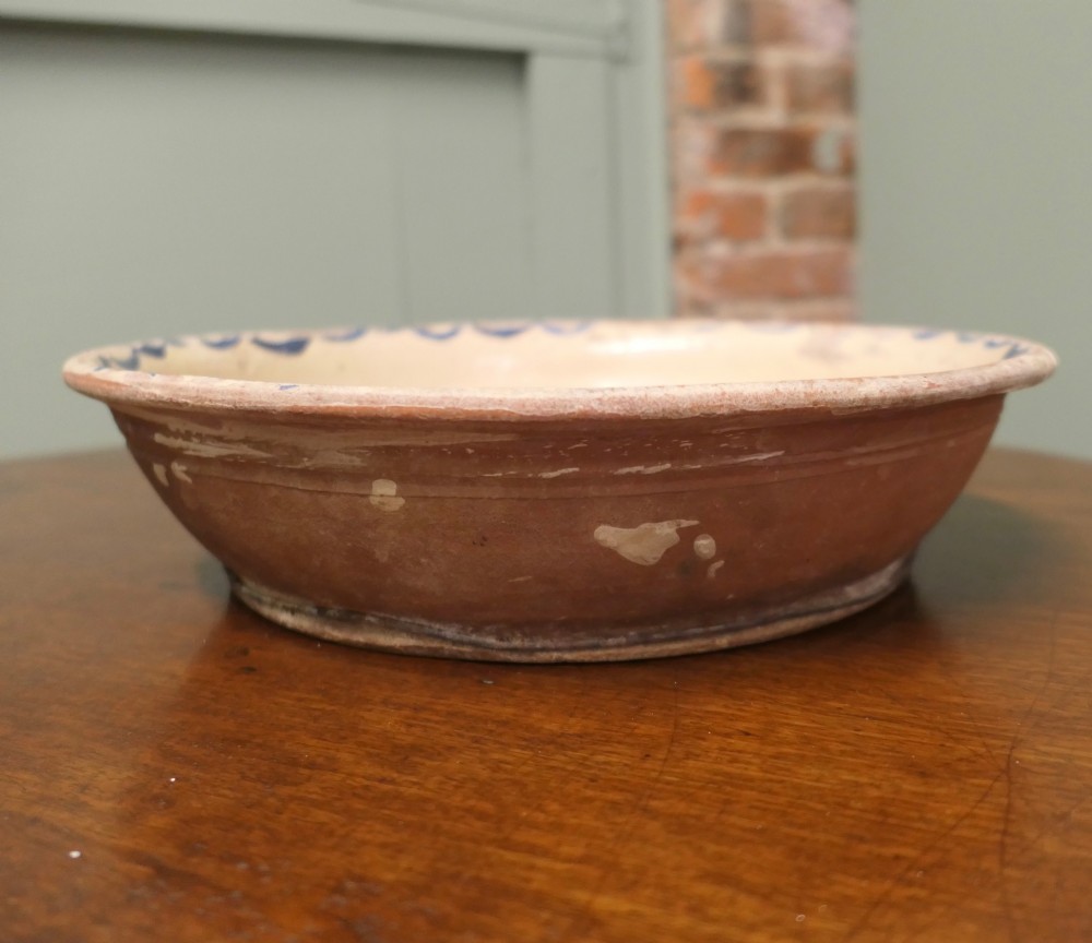 A large round cream-coloured French slipware terracotta bowl decorated with a blue scalloped rim and a naive central illustration of a hunter carrying a bow and hare. View II