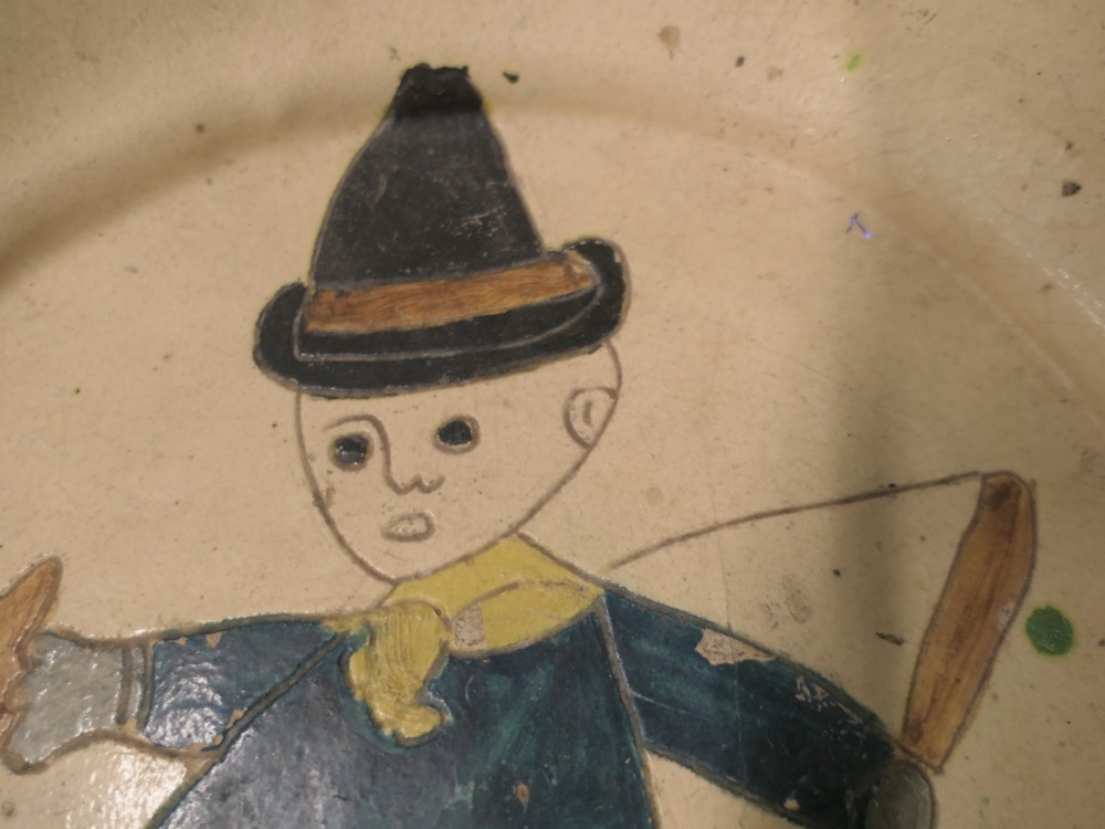 A large round cream-coloured French slipware terracotta bowl decorated with a blue scalloped rim and a naive central illustration of a hunter carrying a bow and hare. View III