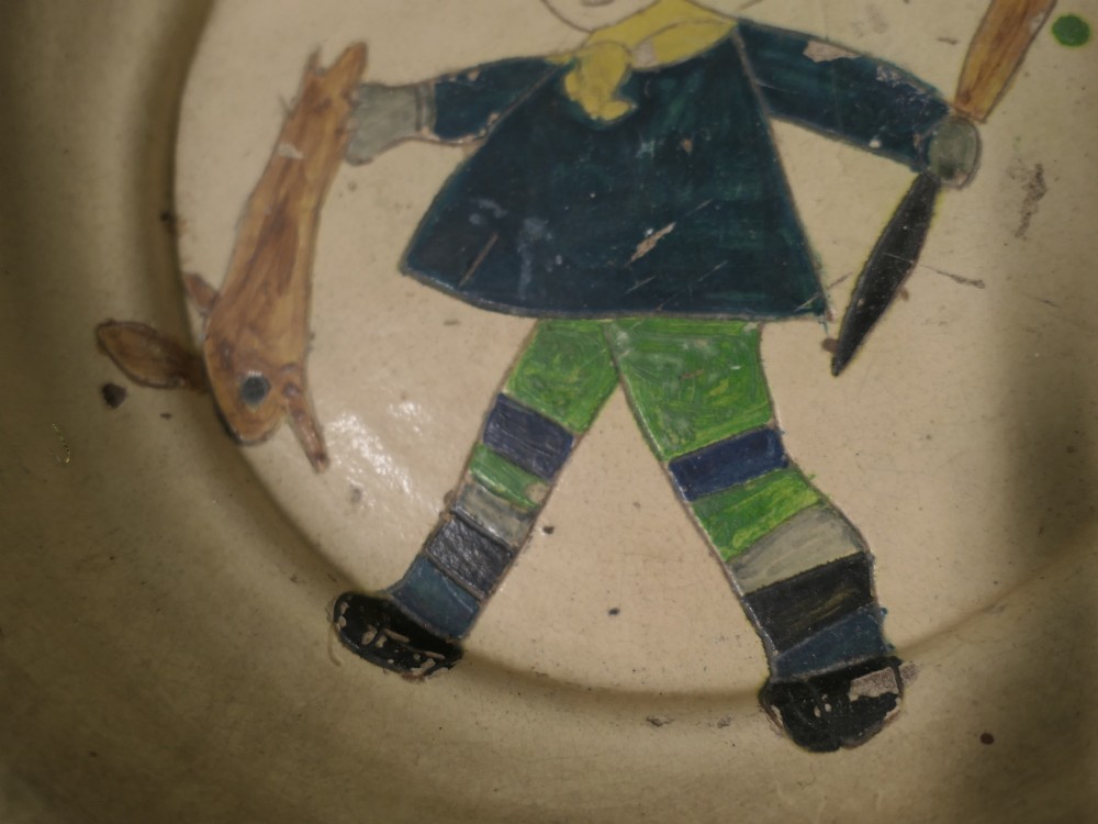 A large round cream-coloured French slipware terracotta bowl decorated with a blue scalloped rim and a naive central illustration of a hunter carrying a bow and hare. View IV