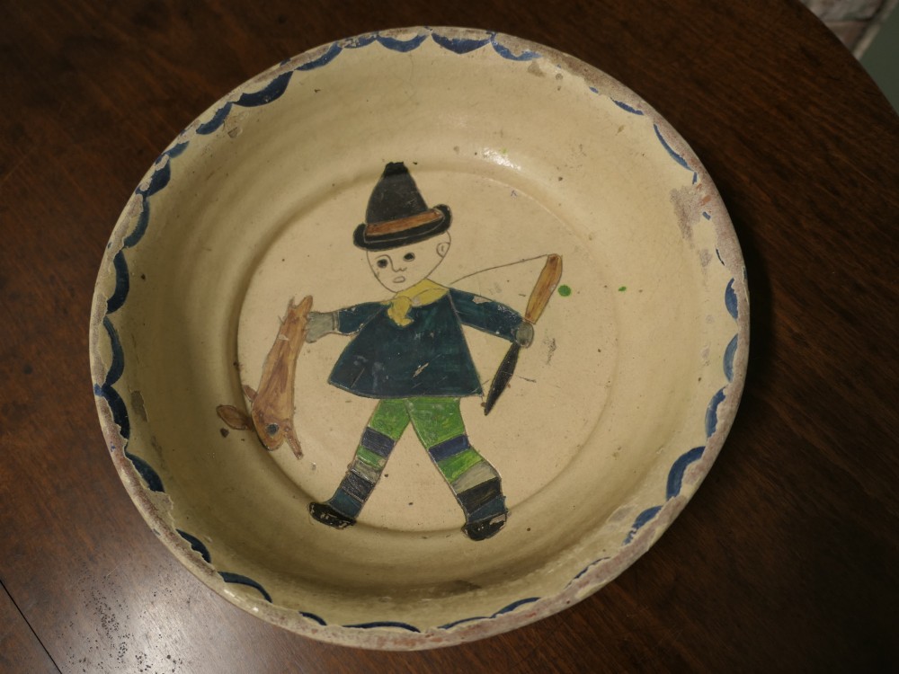 A large round cream-coloured French slipware terracotta bowl decorated with a blue scalloped rim and a naive central illustration of a hunter carrying a bow and hare. View V