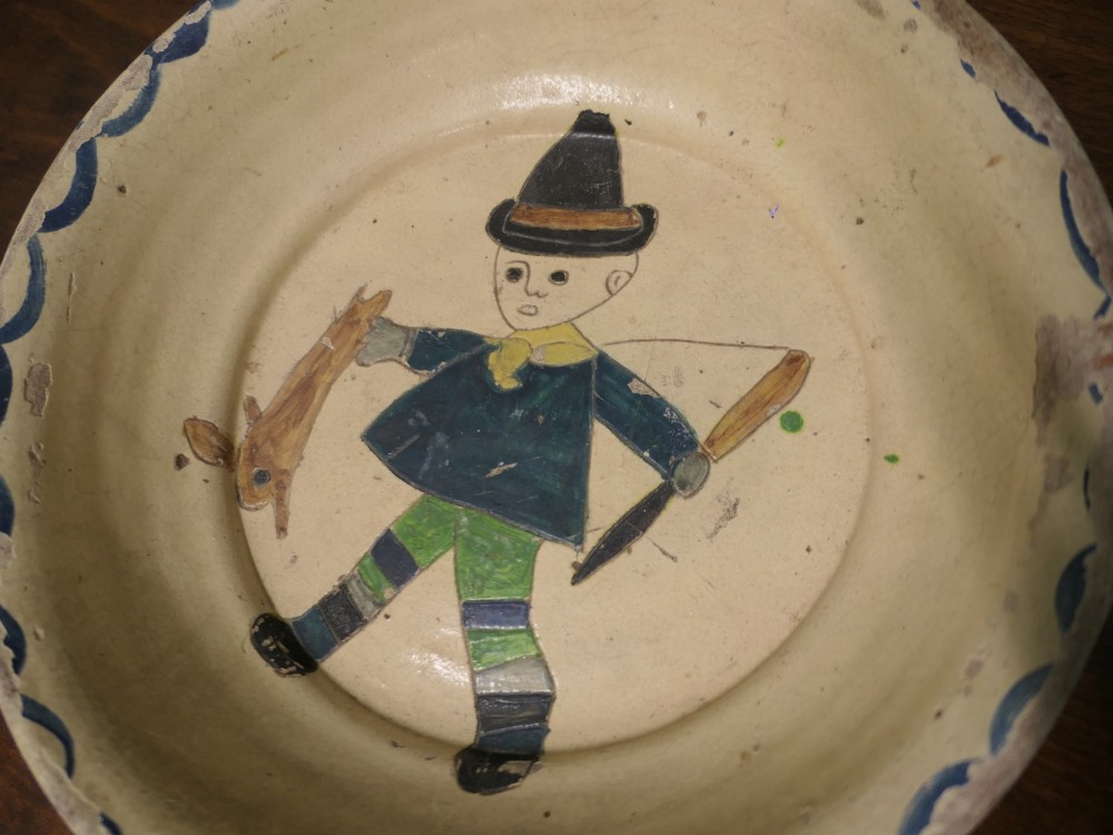 A large round cream-coloured French slipware terracotta bowl decorated with a blue scalloped rim and a naive central illustration of a hunter carrying a bow and hare. View VII