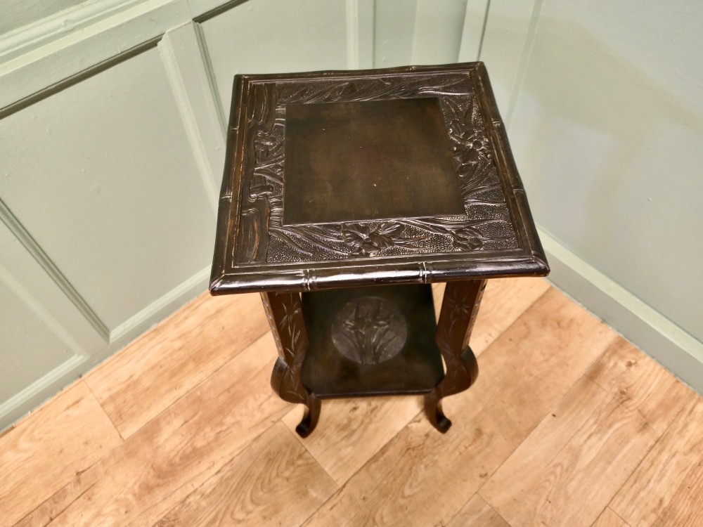 A dark brown Arts and Crafts lamp table featuring carved iris floral motifs, bamboo moulding along the rim, flared lower legs, and an undertier. View II