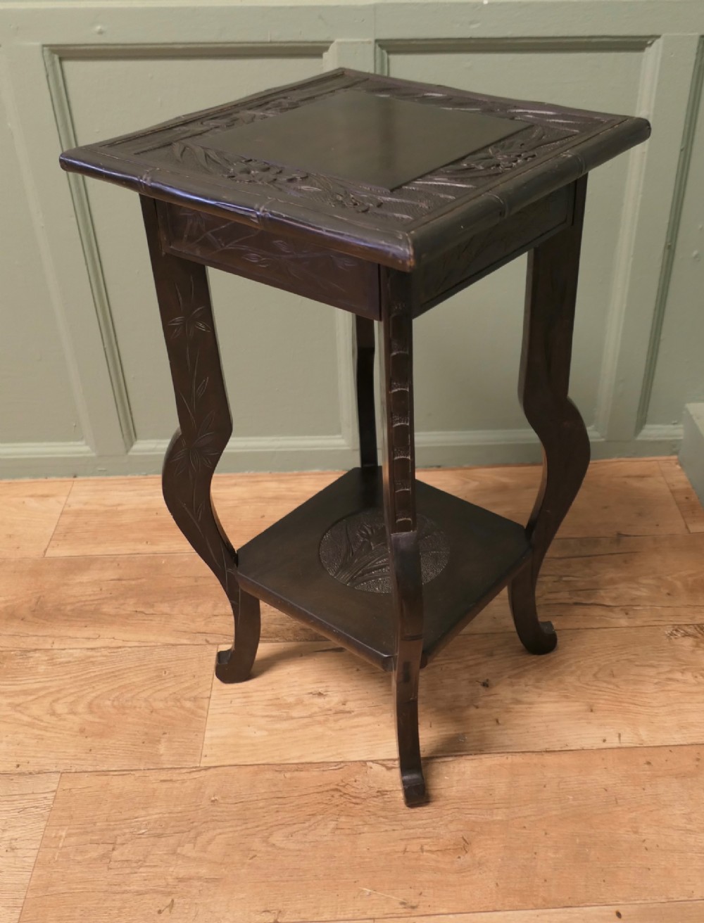 A dark brown Arts and Crafts lamp table featuring carved iris floral motifs, bamboo moulding along the rim, flared lower legs, and an undertier. View III