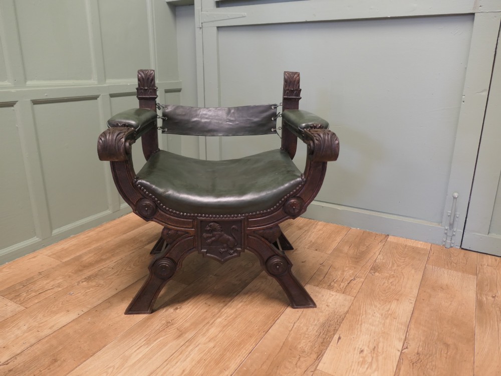 A 19th-century carved oak X-frame armchair featuring green leather upholstery, acanthus finials, and a front crossbar carved with a lion shield.