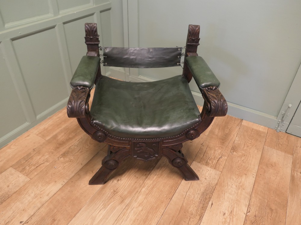 A 19th-century carved oak X-frame armchair featuring green leather upholstery, acanthus finials, and a front crossbar carved with a lion shield. View II