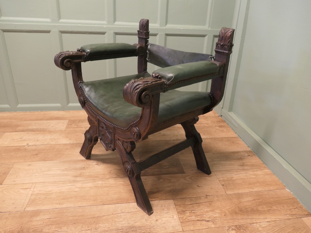 A 19th-century carved oak X-frame armchair featuring green leather upholstery, acanthus finials, and a front crossbar carved with a lion shield. View III