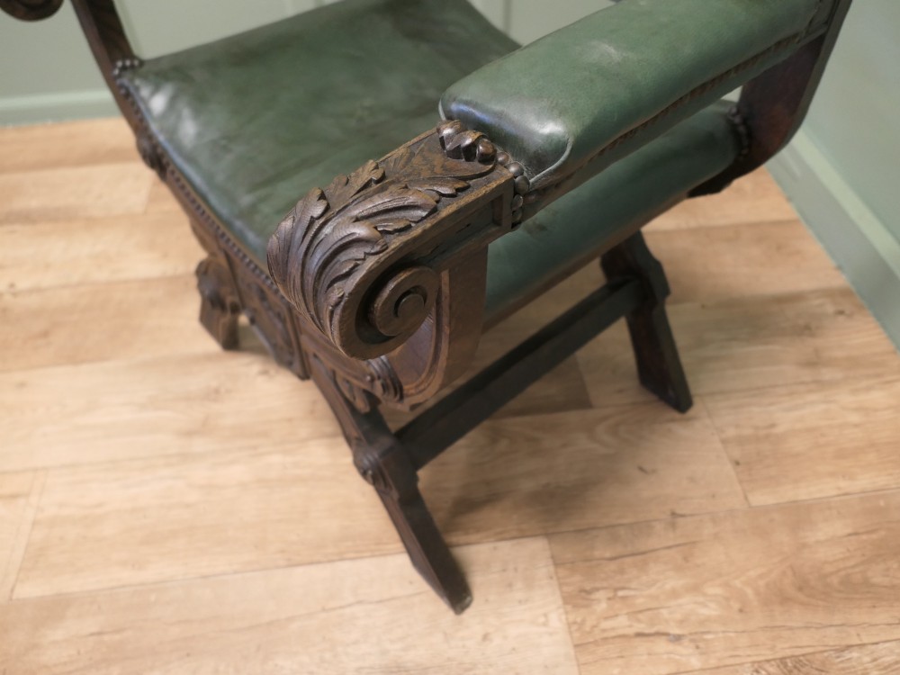 A 19th-century carved oak X-frame armchair featuring green leather upholstery, acanthus finials, and a front crossbar carved with a lion shield. View IV