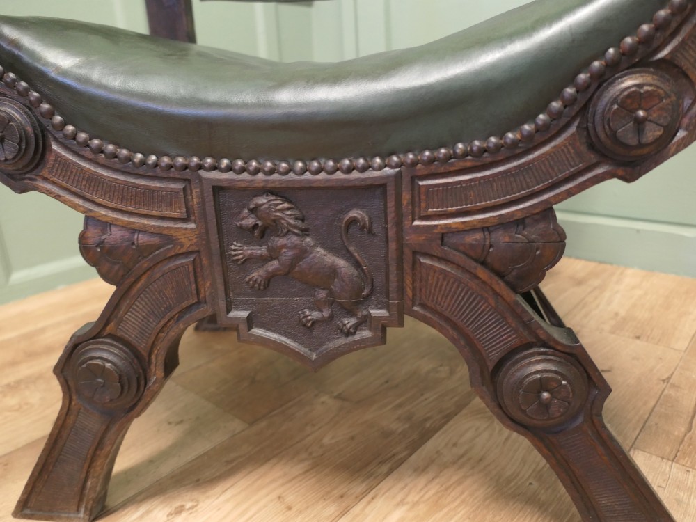 A 19th-century carved oak X-frame armchair featuring green leather upholstery, acanthus finials, and a front crossbar carved with a lion shield. View V