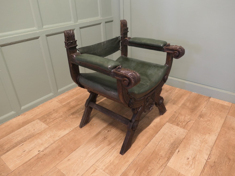 A 19th-century carved oak X-frame armchair featuring green leather upholstery, acanthus finials, and a front crossbar carved with a lion shield. View VI