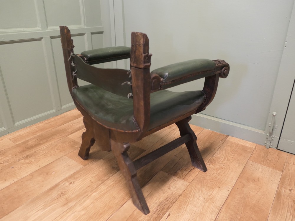 A 19th-century carved oak X-frame armchair featuring green leather upholstery, acanthus finials, and a front crossbar carved with a lion shield. View VII